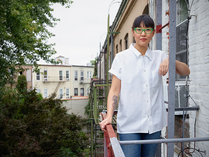 Chef Kate Telfeyan stands on a fire escape with green-rimmed glasses.