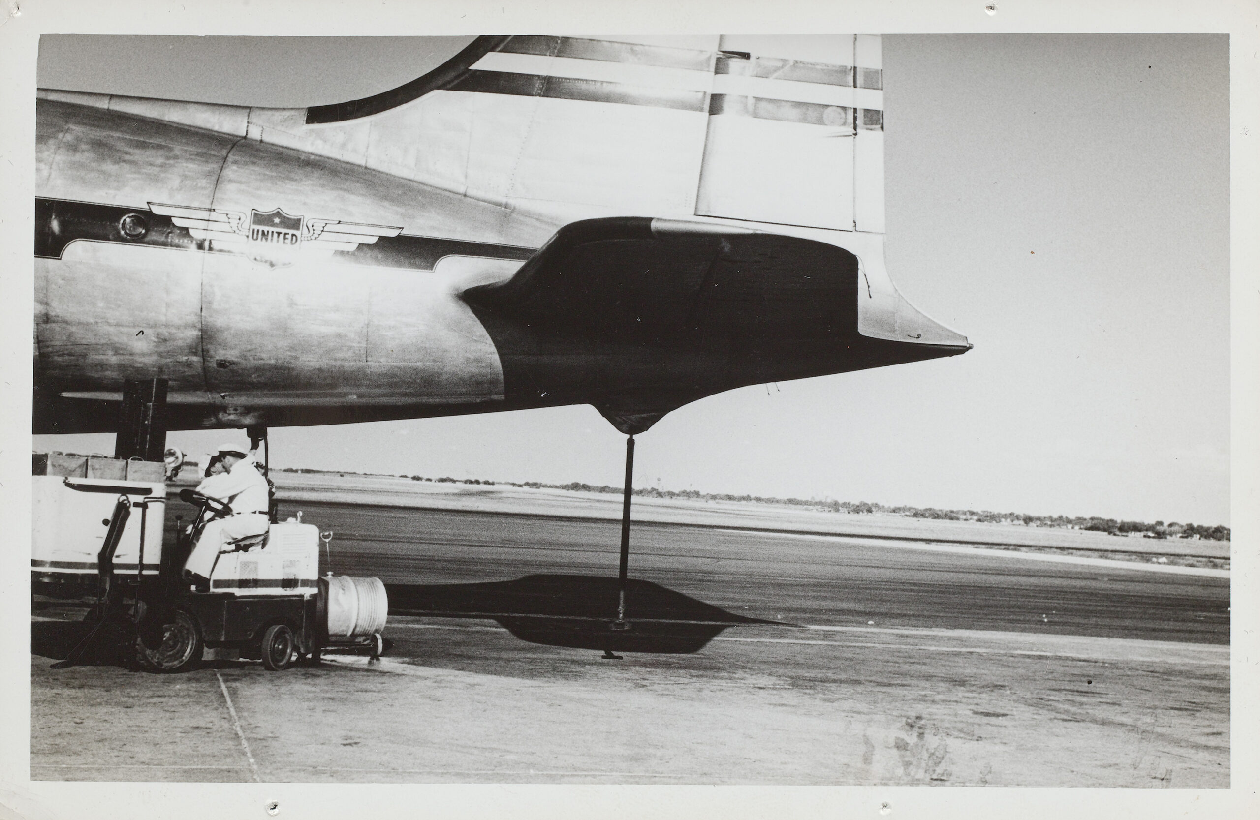 Black and white photograph of a plane tail on a tarmac with two figures in cart under the plane.