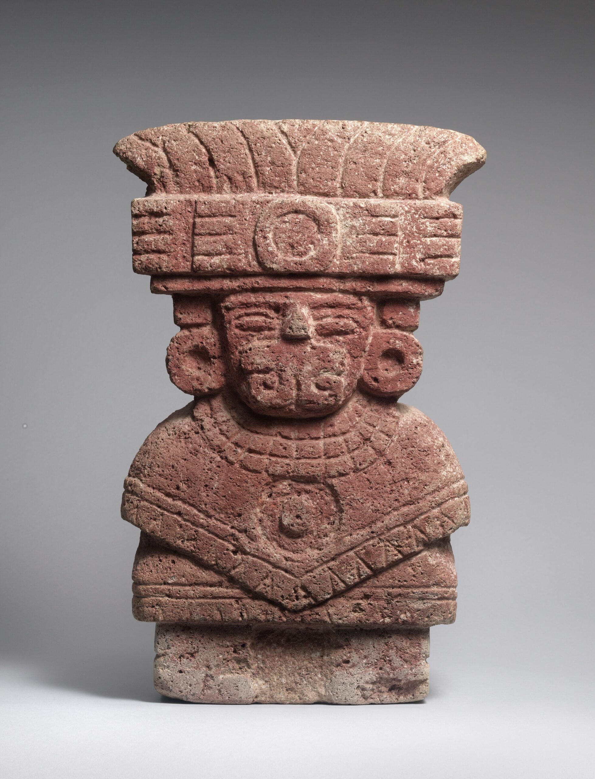 Seated stone figure with an elaborate headdress, garment, and ear spools.