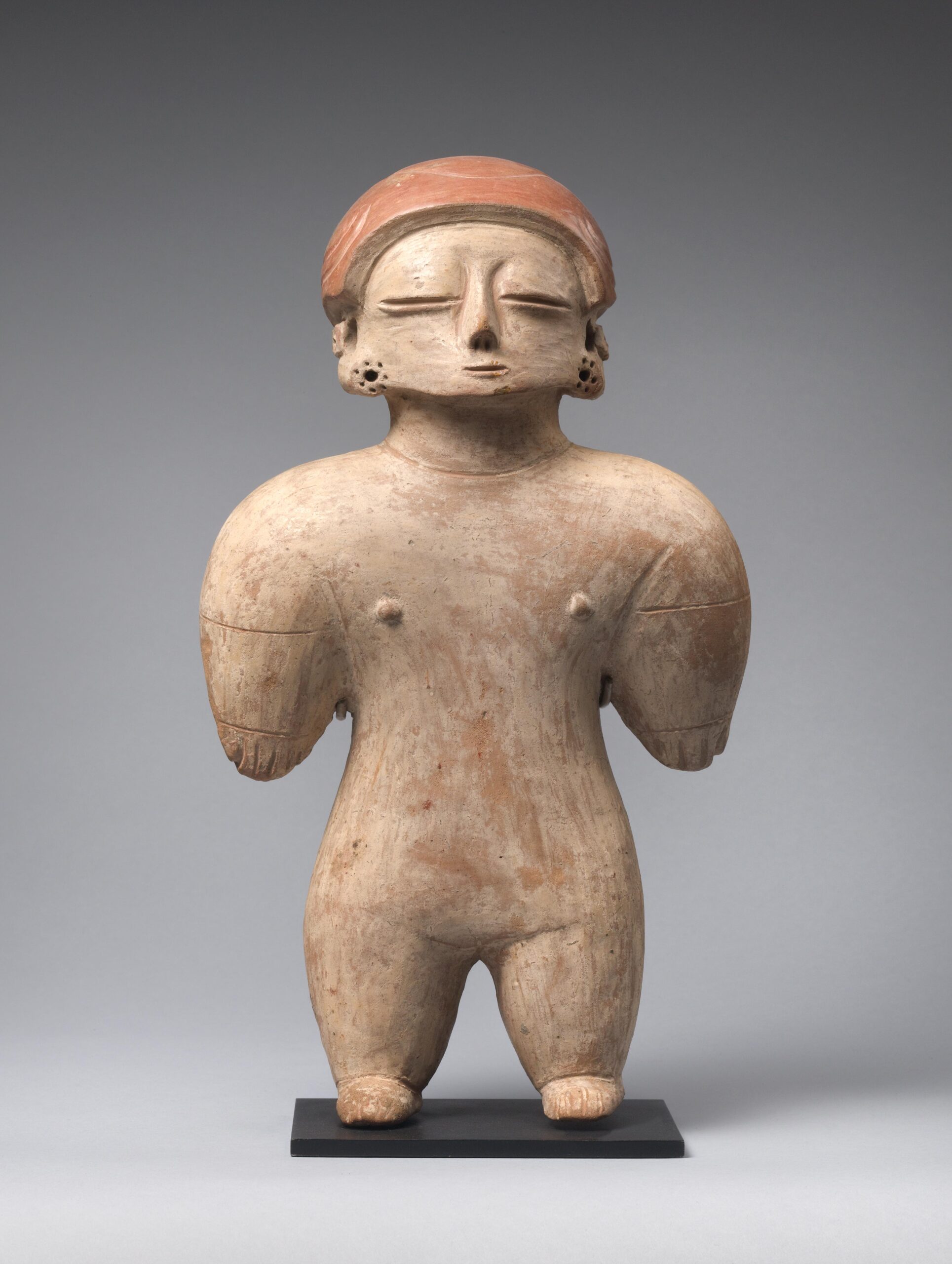 Stylized ceramic standing female figure with short round shoulders and legs, a headress, and ear spools.