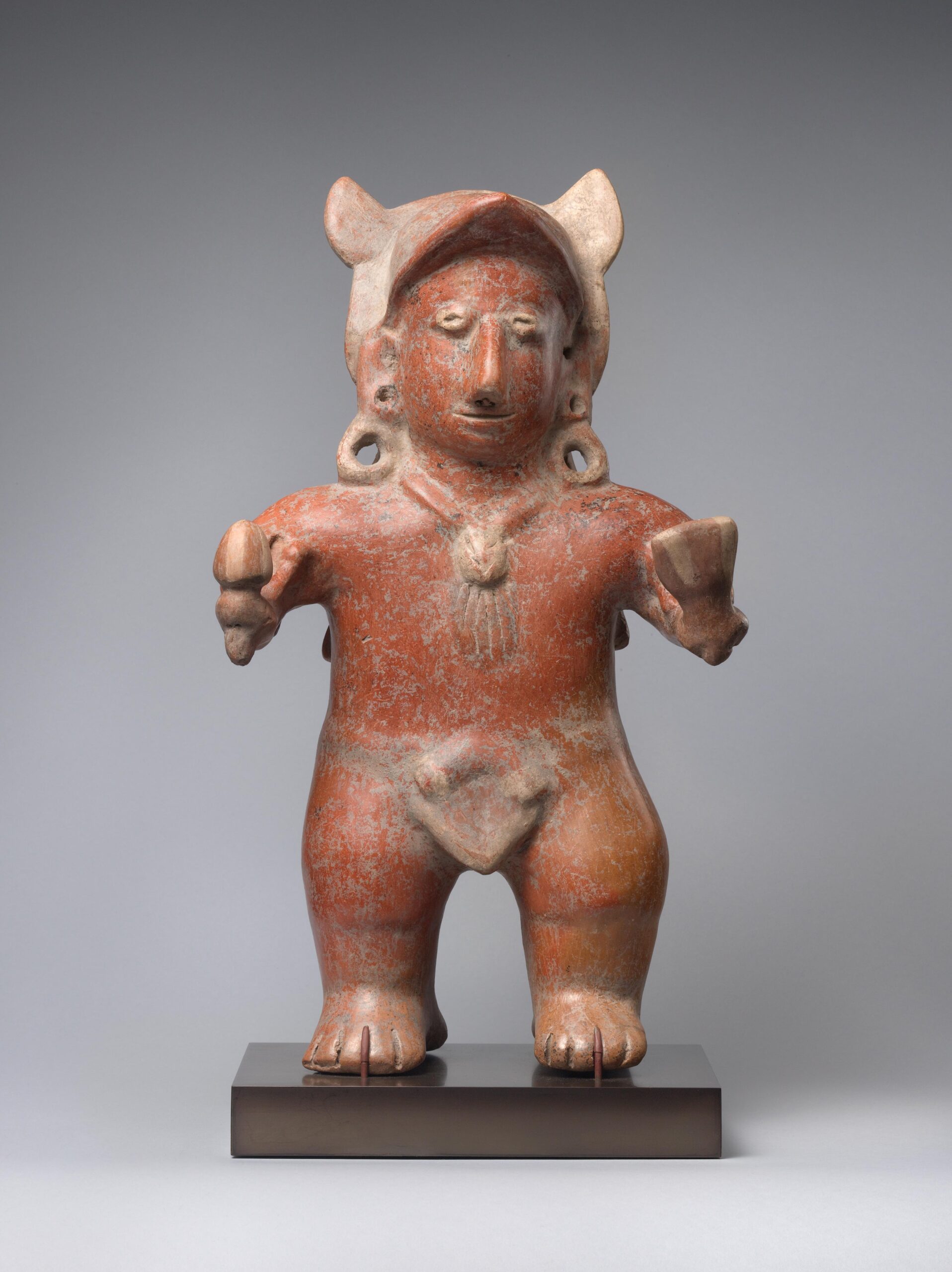 A ceramic sculpture of a red figure wearing an animal head, loin cloth, and earrings, holding a rattle and a fan.