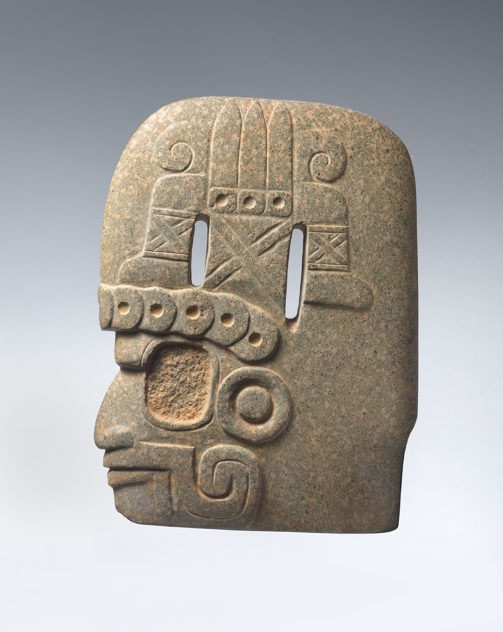Flat stone carving of human face shown in profile with elaborate headpiece.
