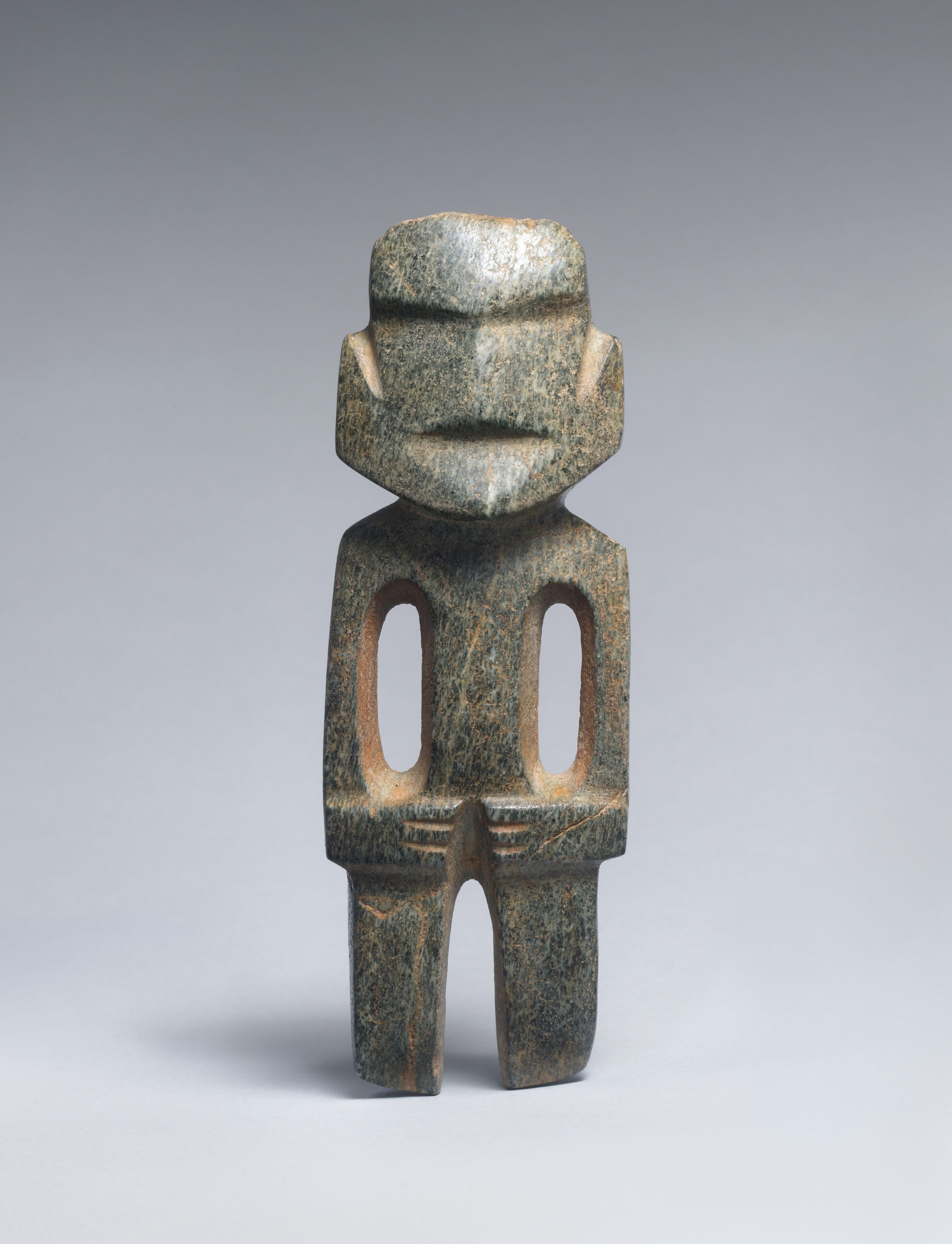 Stone figure with a broad rounded face, indentations for the nose and mouth, two hollows to form the torso and arms, and hands folded across the body.