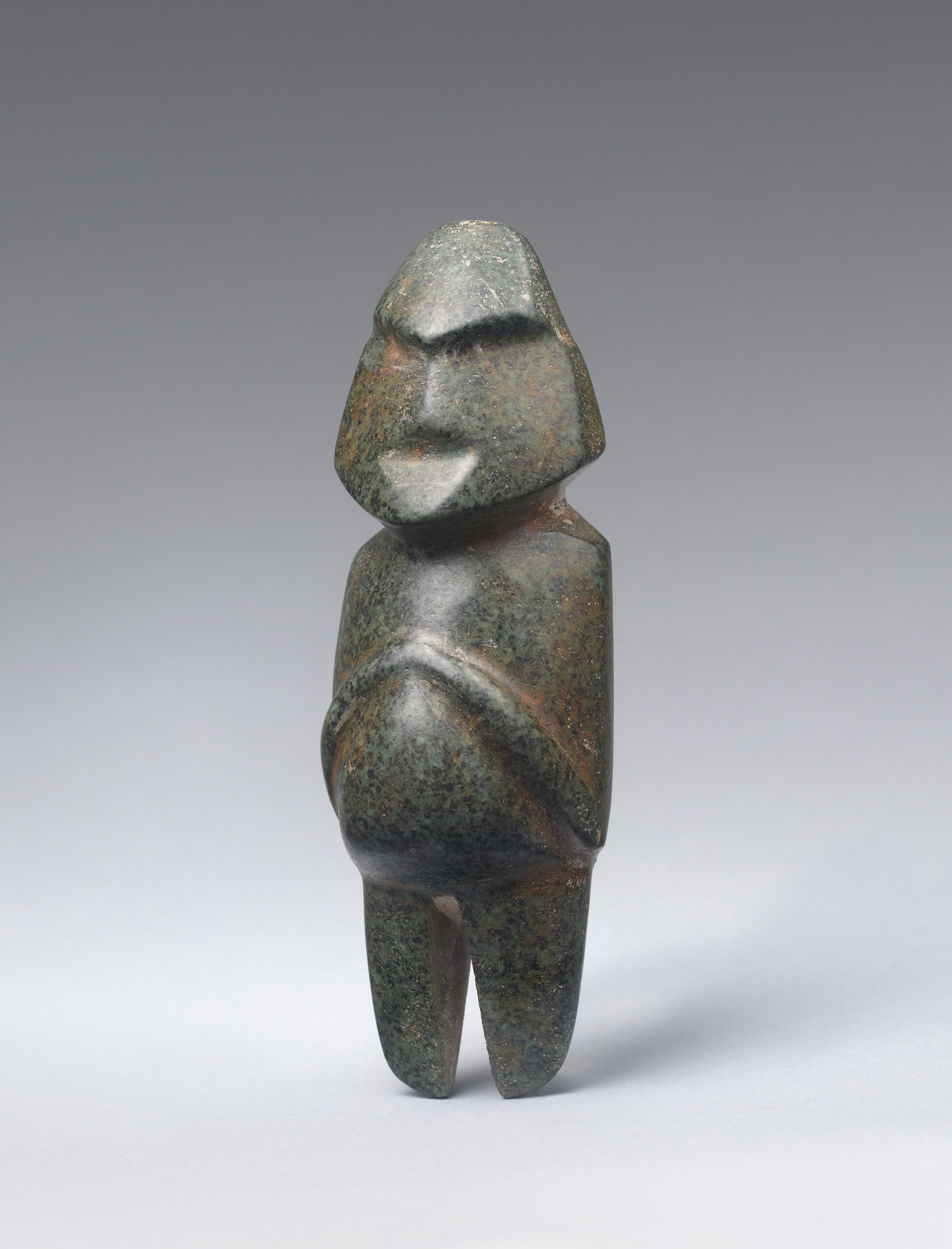 Small, abstract stone carving of a standing human figure with broad, pointed facial features and folded hands upon its round abdomen.