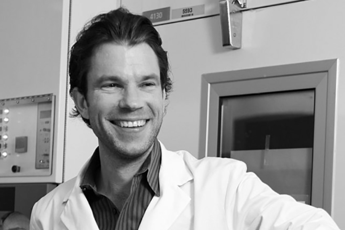 A black and white photo of Konrad Hochedlinger wearing a lab coat in front of equipment.