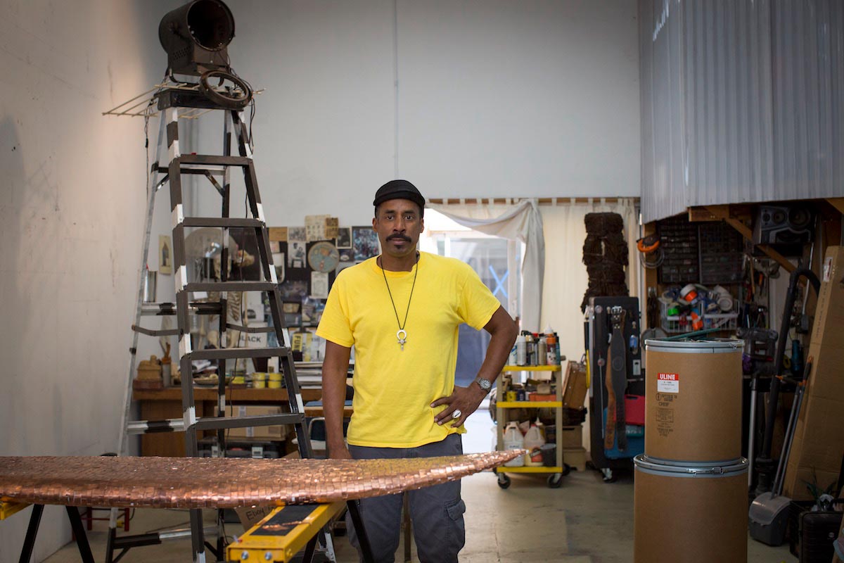 Nari Ward stands in his art studio surrounded by supplies and equipment, including a tall ladder and a bronze surfboard.