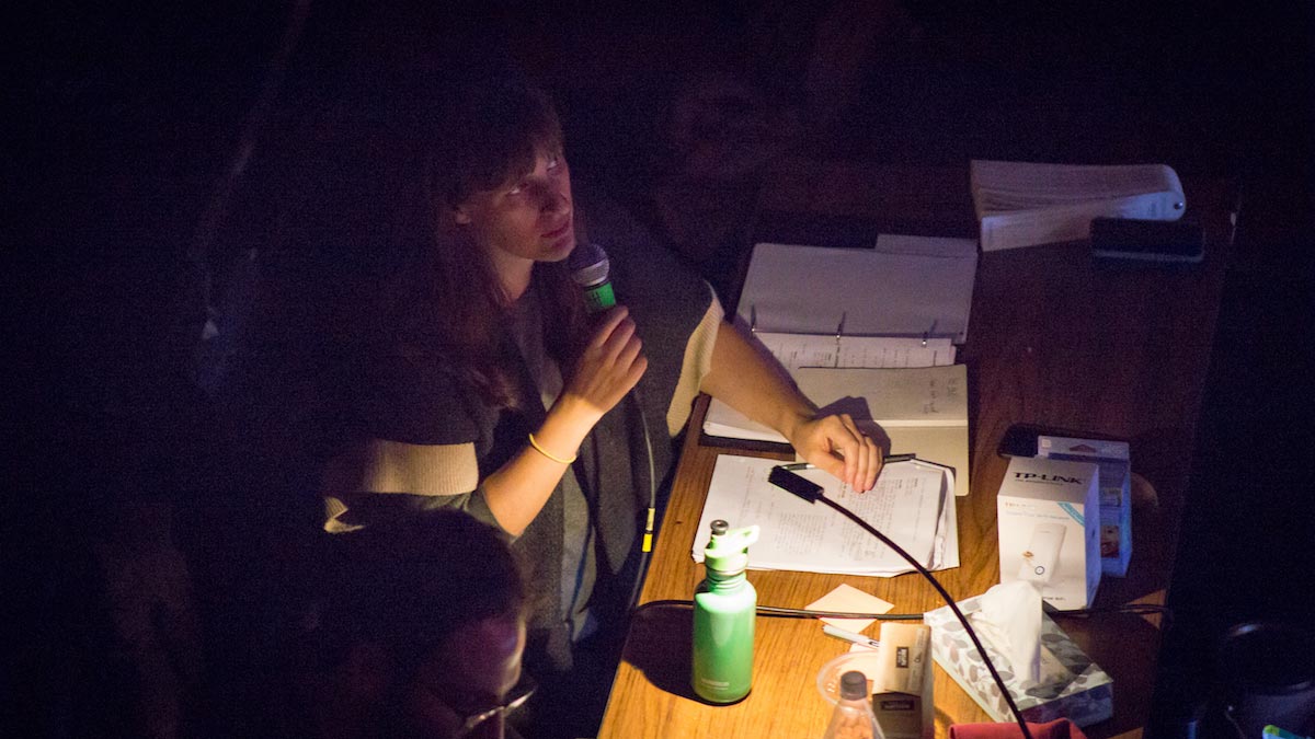 Sarah Benson is sitting in a dark, isolated part of a theater lit by a small desk-lamp, holding a microphone and a pen with scripts in front of her.