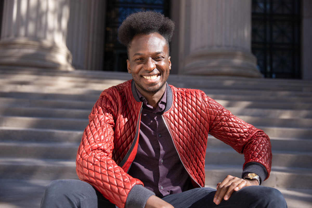 Ibrahim Cissé wearing a red jacket sits smiling on the steps of an MIT building.
