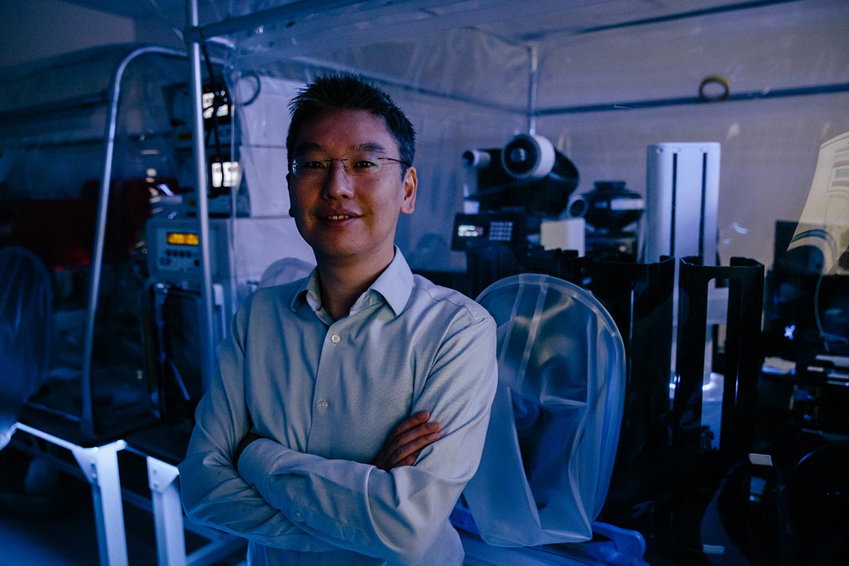 Harris Wang, arms folded, stands in a dark room in front of a microscope.