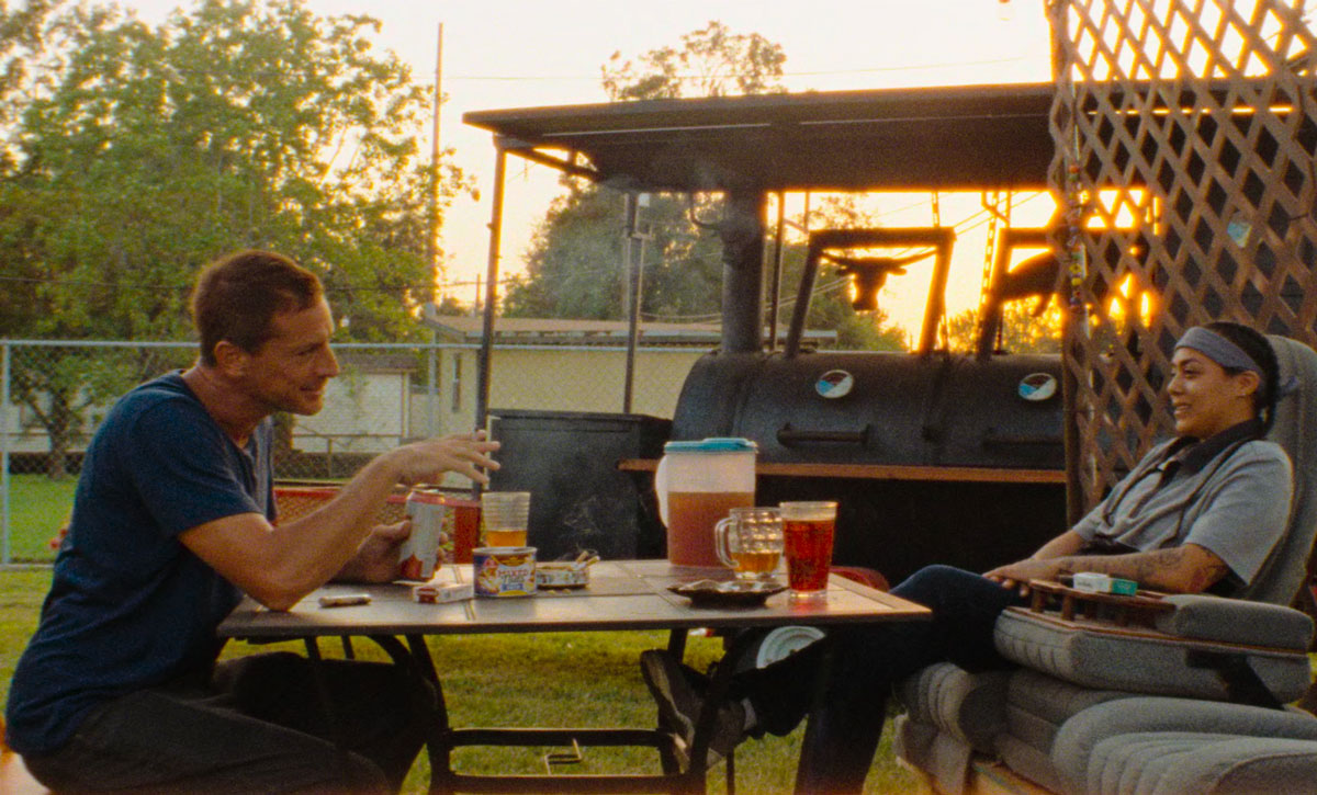 Two characters from 'Red Rocket' sit at a garden table drinking iced tea. 