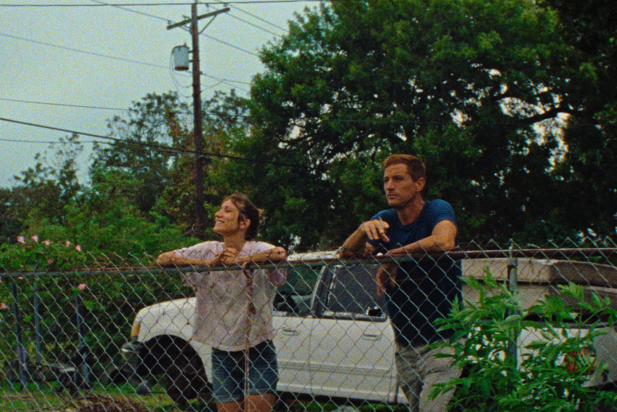Two characters from 'Red Rocket' lean against a wire fence with a car and green trees behind them.