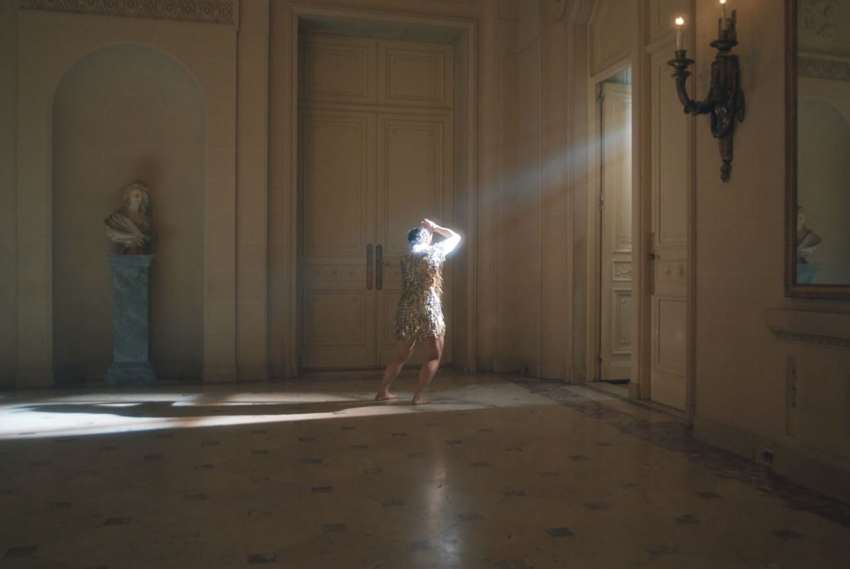A dancer stands in a light beam with their arm raised in front of their face.