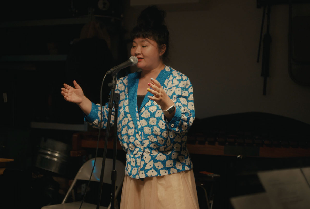 Du Yun dressed in a blue and white floral blazer and singing at a microphone. 