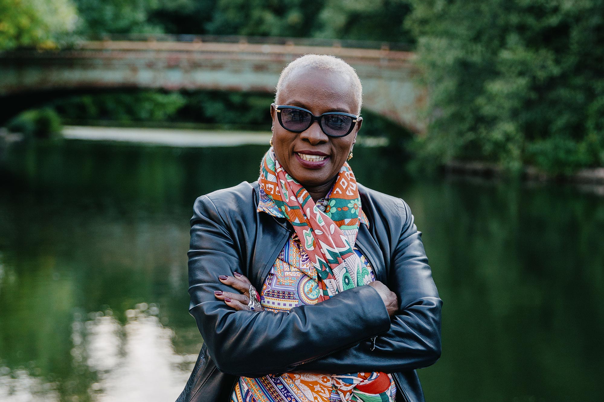 Angélique Kidjo smiling, arms crossed, in front of a lake and a bridge.